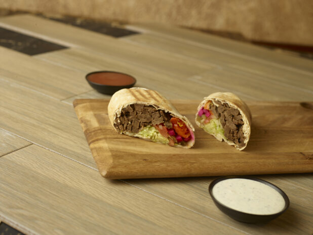 Beef shawarma on a wooden board cut in half with dips on the side on a 45 degree angle