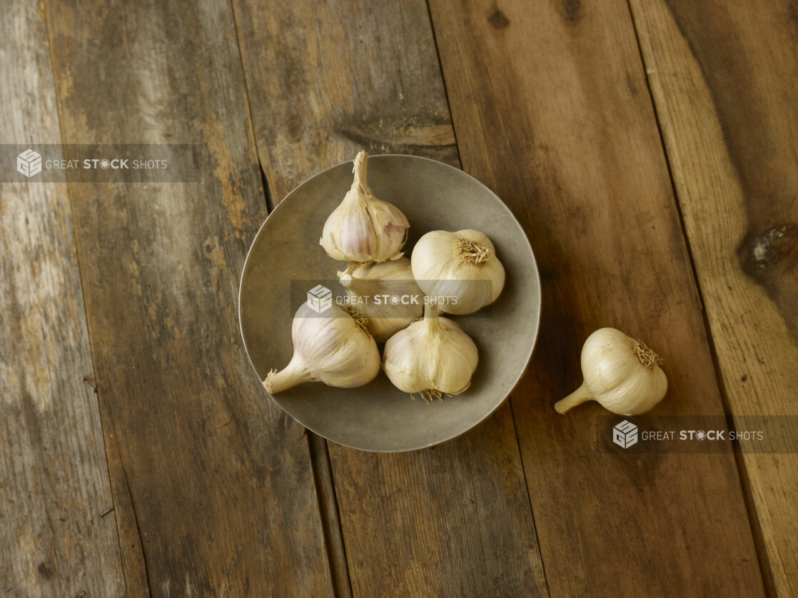 5 garlic bulbs in a bowl on a aged wooden table with an overhead view with 1 garlic bulb to the right of the bowl