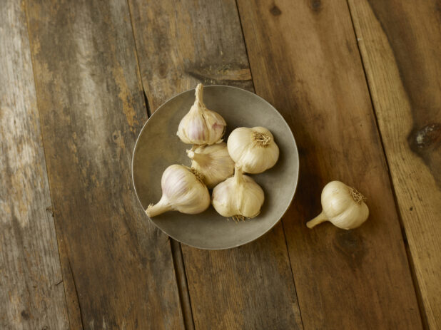 5 garlic bulbs in a bowl on a aged wooden table with an overhead view with 1 garlic bulb to the right of the bowl