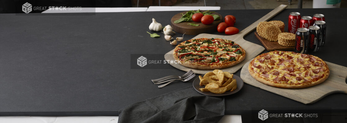 2-3 topping pizzas on wooden peels with coke and coke zero, pizzelle, fresh garlic, roma tomatoes, basil, black napkin and forks all on a black background shot left of centre