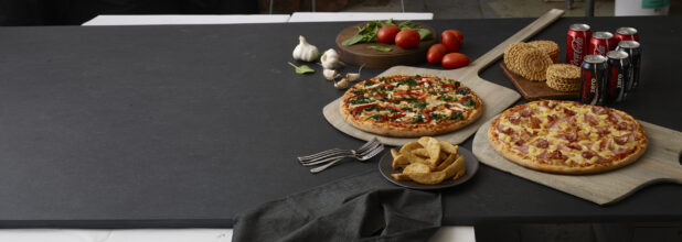 2-3 topping pizzas on wooden peels with coke and coke zero, pizzelle, fresh garlic, roma tomatoes, basil, black napkin and forks all on a black background shot left of centre