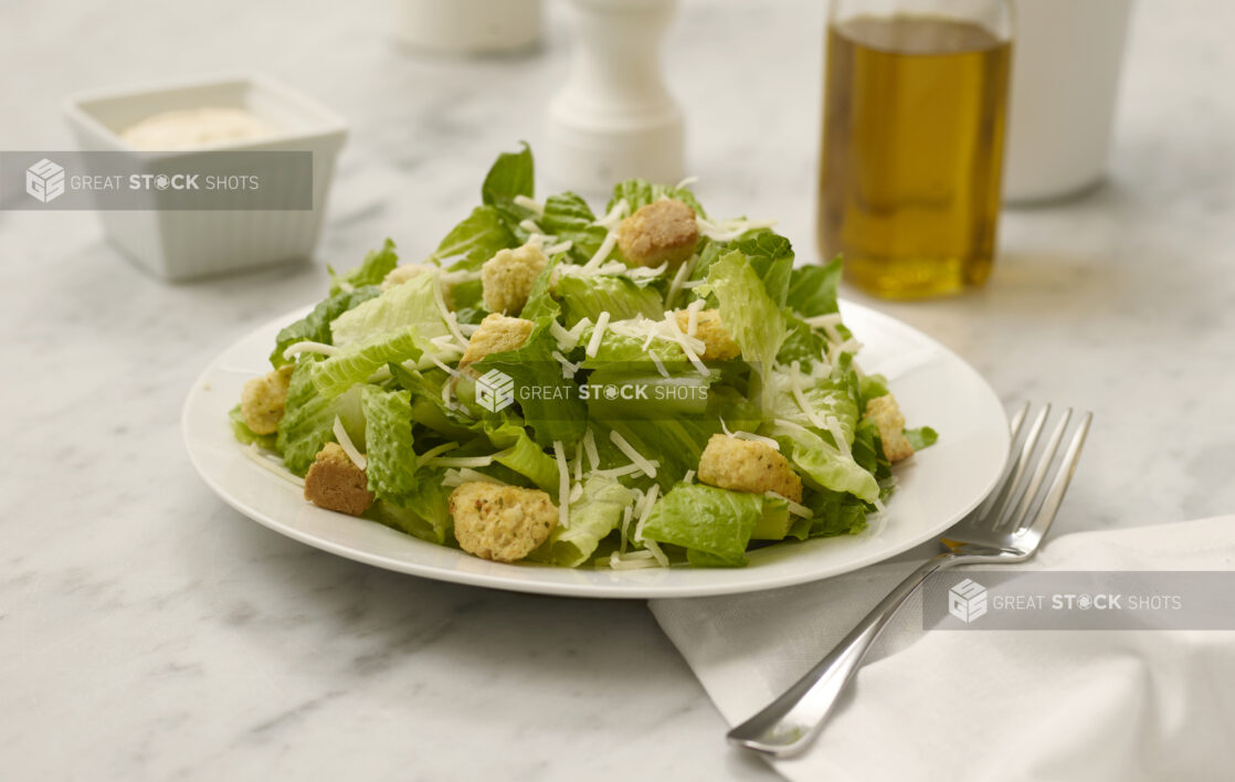 Caesar salad in a white bowl on a white marble table with olive oil and dressing in the background