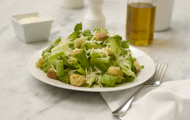 Caesar salad in a white bowl on a white marble table with olive oil and dressing in the background