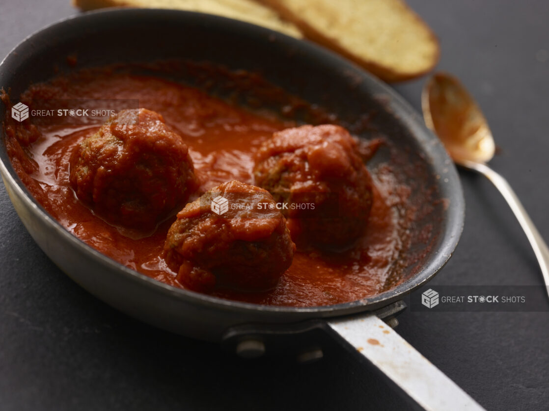 3 meatballs in a pan with tomato sauce in close up shot on an angle with a spoon and garlic bread in the background all on a black background