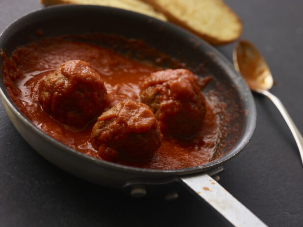 3 meatballs in a pan with tomato sauce in close up shot on an angle with a spoon and garlic bread in the background all on a black background