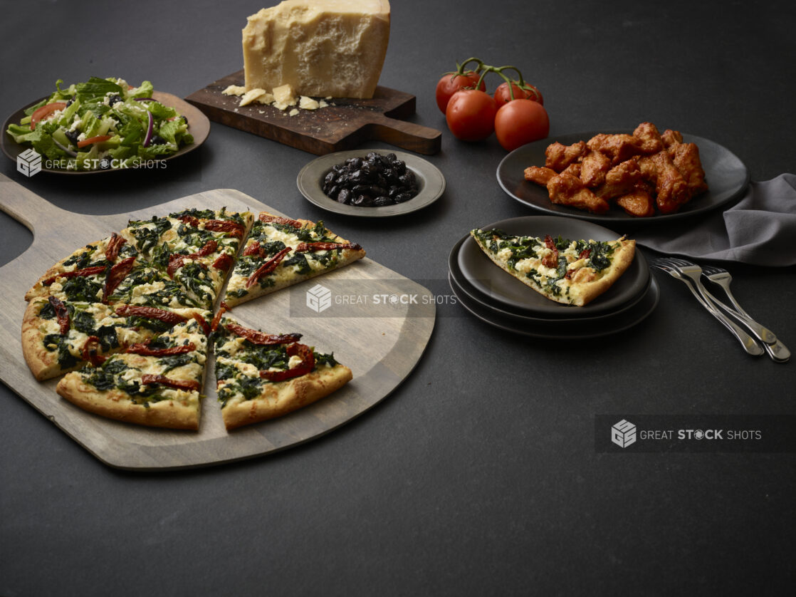 3 topping Mediterranean-style pizza, sauced chicken wings, side salad, whole parmesan and a side bowl of black olives on a black background on a 45 degree angle