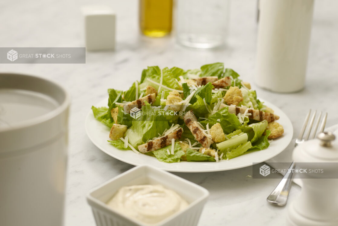 Chicken Caesar salad in a white bowl on a white marble table with dressing in the foreground