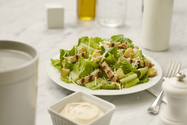 Chicken Caesar salad in a white bowl on a white marble table with dressing in the foreground