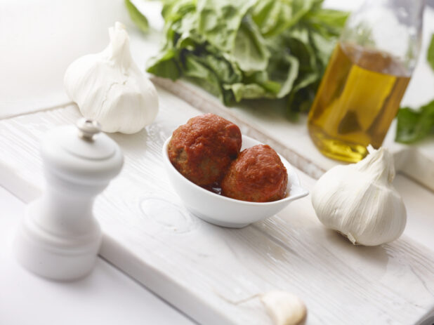 2 meatballs with tomato sauce in a ramekin on a white board surrounded by a white pepper mill, whole garlic bulbs, fresh basil and olive oil all on a white background shot on an angle