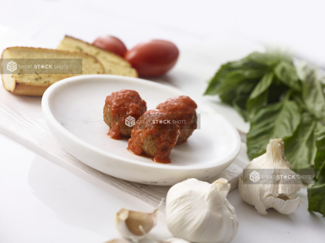 3 meatballs with tomato sauce on a white side plate surrounded by fresh basil, garlic, tomatoes and garlic bread on a white table shot on an angle