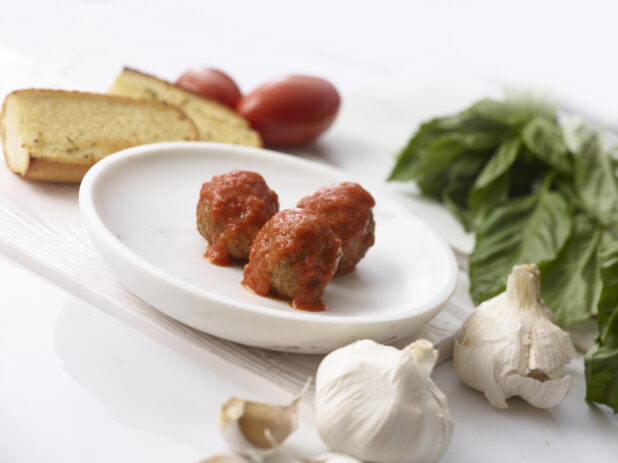 3 meatballs with tomato sauce on a white side plate surrounded by fresh basil, garlic, tomatoes and garlic bread on a white table shot on an angle