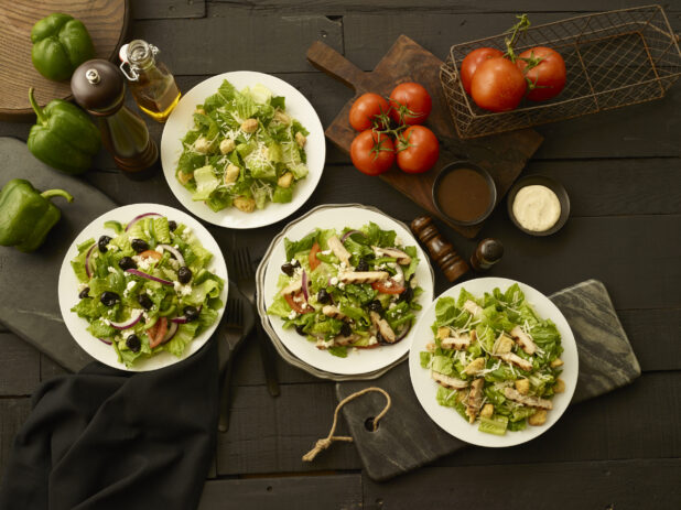 Greek salads and Caesar salads in white bowls on a dark wooden background, with fresh whole vegetables and condiments, overhead shot