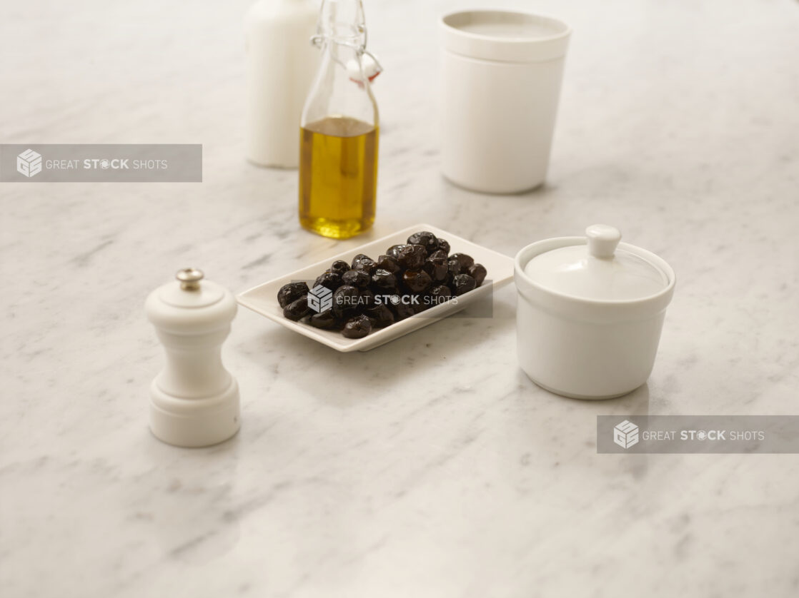 Black olives on a white rectangle plate on a white marble table with olive oil in a bottle and a white pepper mill, 45 degree angle