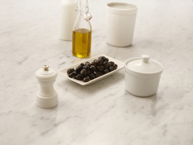 Black olives on a white rectangle plate on a white marble table with olive oil in a bottle and a white pepper mill, 45 degree angle