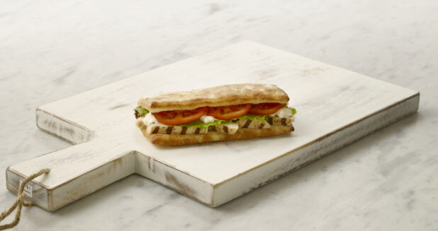 Whole grilled chicken sandwich/hoagie on a white cutting board on a white marble table