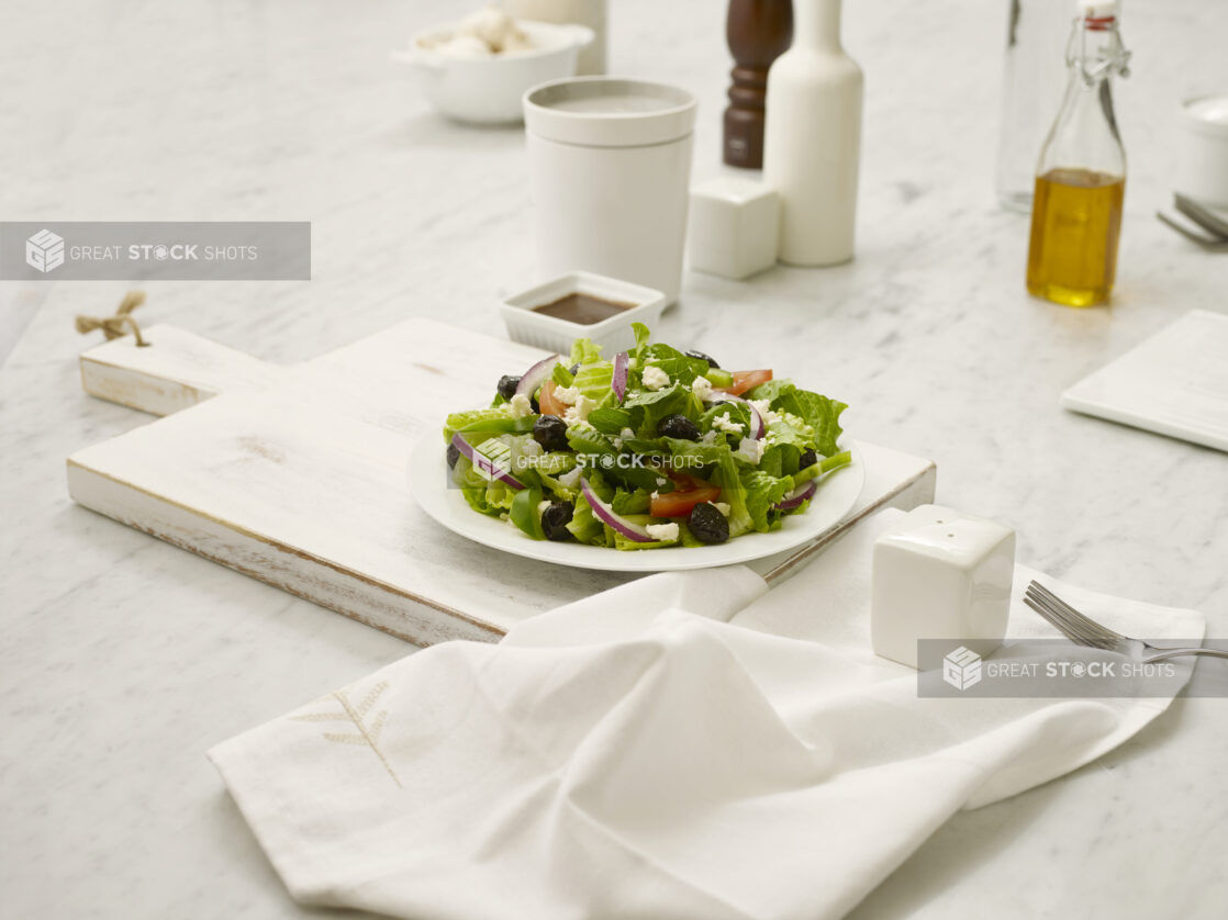 Side greek salad on a white plate on a white cutting board with a white napkin, fork and white salt shaker all in the foreground on a marble table