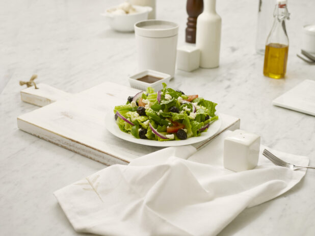 Side greek salad on a white plate on a white cutting board with a white napkin, fork and white salt shaker all in the foreground on a marble table