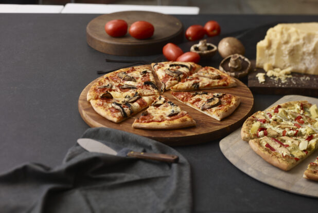 2 gluten free 2 topping pizzas on wooden boards, with Roma tomatoes, mushrooms, parmesan cheese in the background with a black napkin and pie lifter on a black background