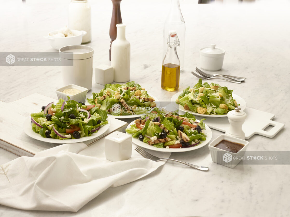 Greek salad and Caesar salad, both with grilled chicken and without, in white bowls on a white marble table with dressing, olive oil in a bottle and other accompaniments, overhead view