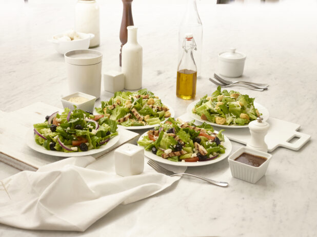 Greek salad and Caesar salad, both with grilled chicken and without, in white bowls on a white marble table with dressing, olive oil in a bottle and other accompaniments, overhead view