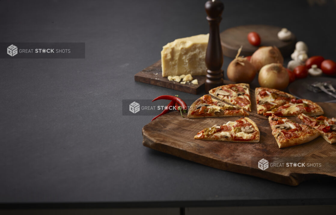 3 topping pizza on a rustic wooden board surrounded by a peppermill, onions, parmesan cheese, and other ingredients on a black background, shot right of centre