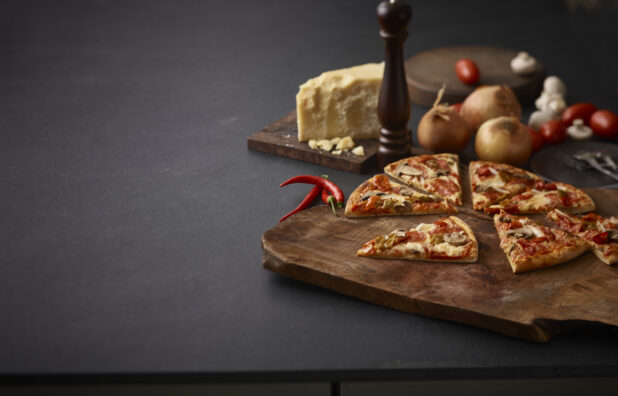 3 topping pizza on a rustic wooden board surrounded by a peppermill, onions, parmesan cheese, and other ingredients on a black background, shot right of centre