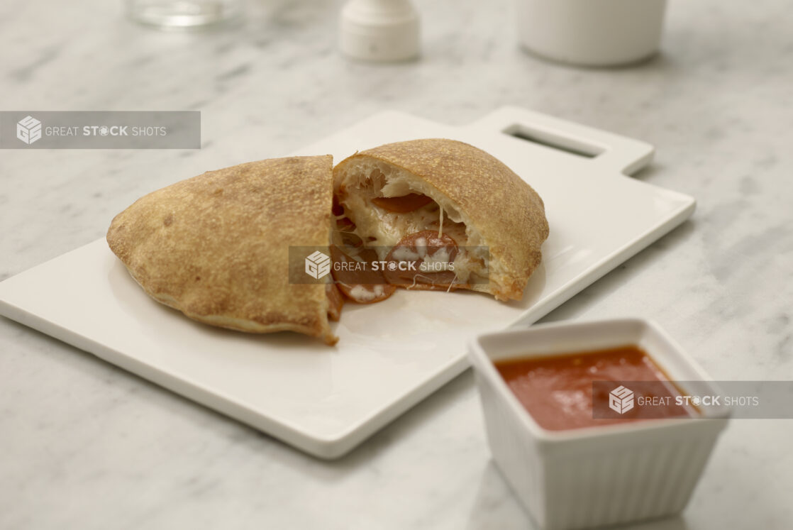 A calzone cut in half on a white cutting board with a ramekin of pizza sauce  on a white marble table