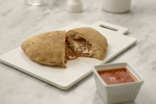 A calzone cut in half on a white cutting board with a ramekin of pizza sauce  on a white marble table