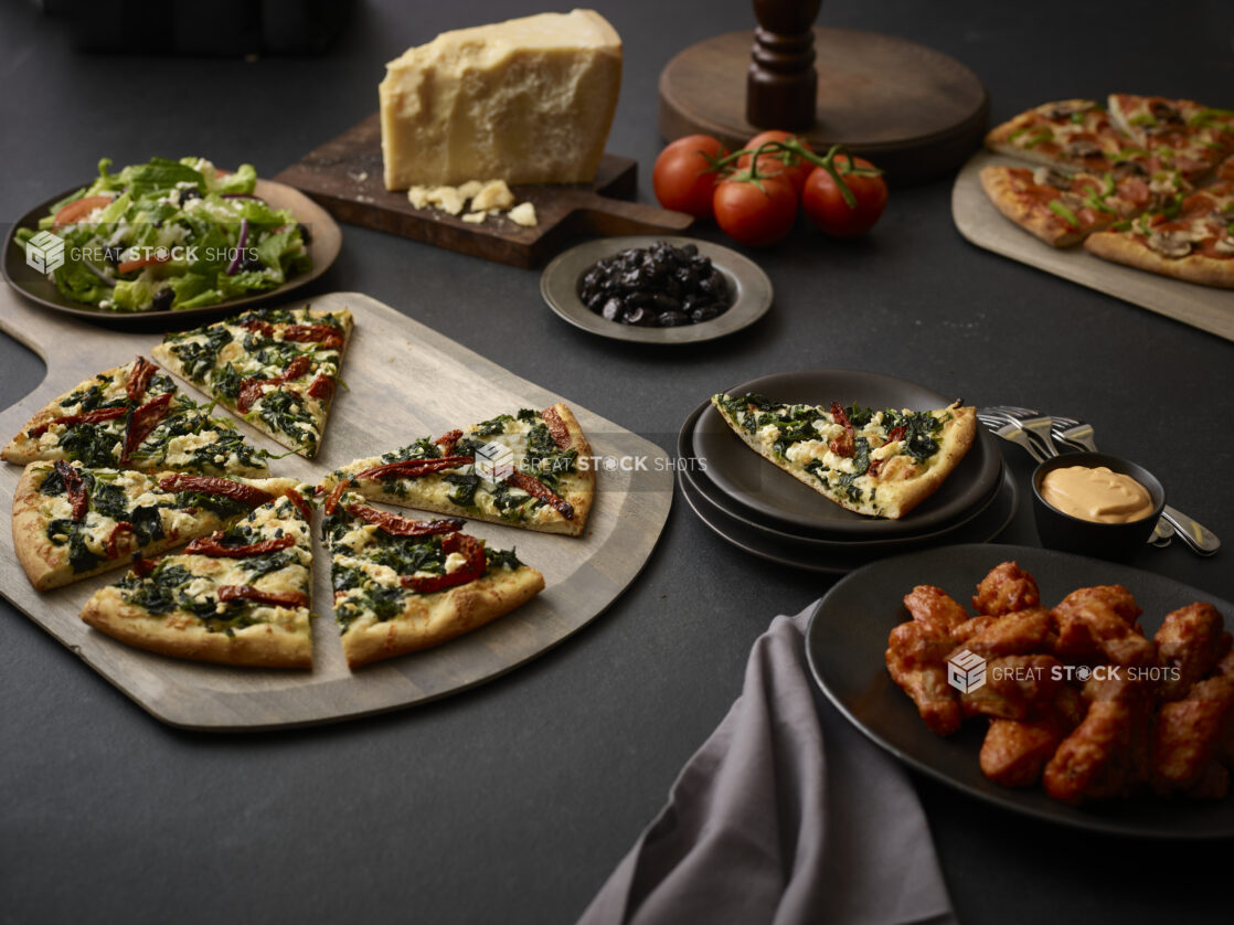 Sliced 2 topping pizza on a wooden cutting board, with various side dishes and fresh ingredients in background, black background