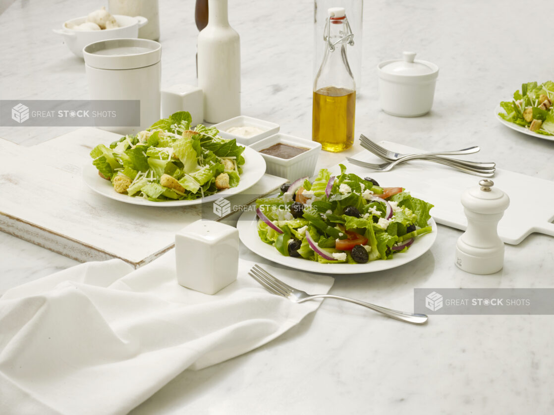 Greek salad and Caesar salad in white bowls on a white marble table with dressing, olive oil in a bottle and other accompaniments