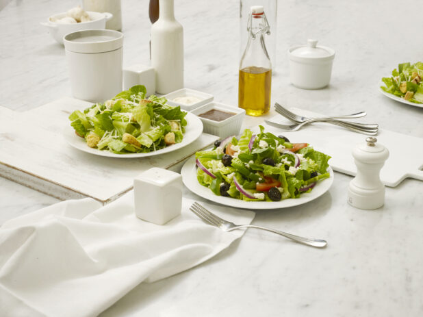 Greek salad and Caesar salad in white bowls on a white marble table with dressing, olive oil in a bottle and other accompaniments