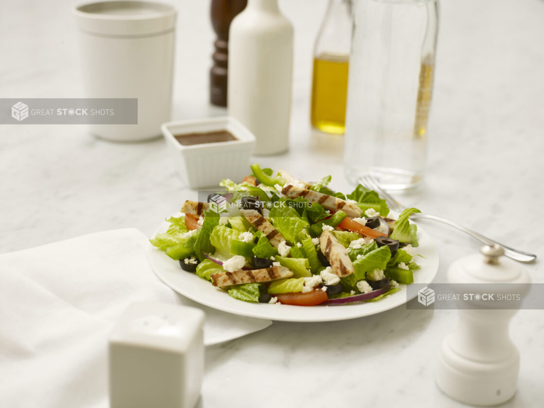 Chicken Caesar salad in a white bowl on a white marble table with a white salt shaker and white pepper mill, a white napkin, a fork, and table accompaniments