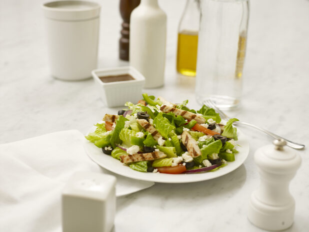 Chicken Caesar salad in a white bowl on a white marble table with a white salt shaker and white pepper mill, a white napkin, a fork, and table accompaniments