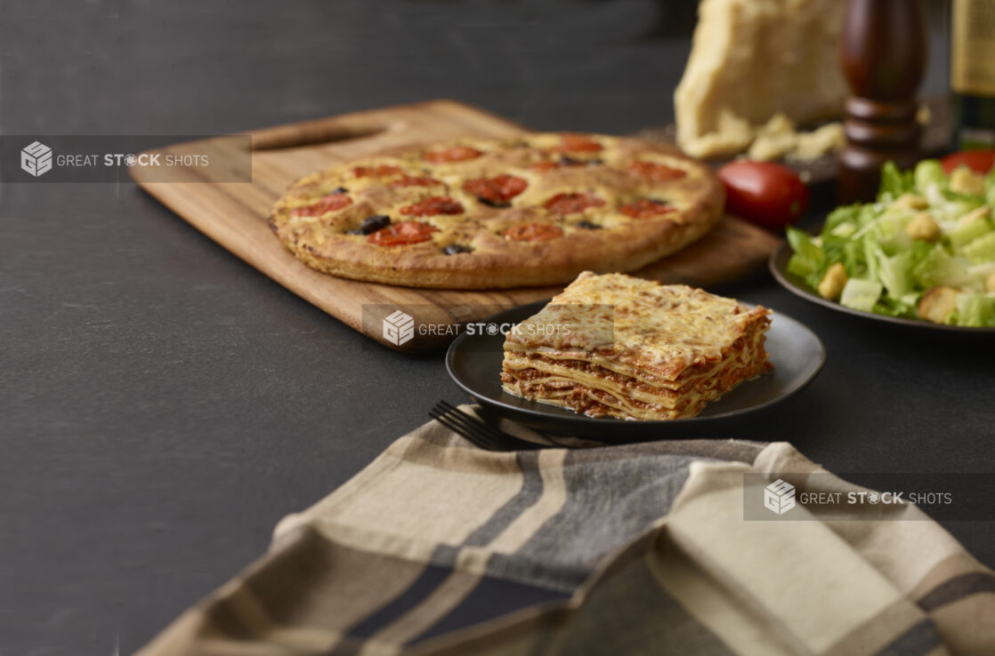 Slice of lasagna on a round black plate, focaccia, side salad, and other table items in background, dark background