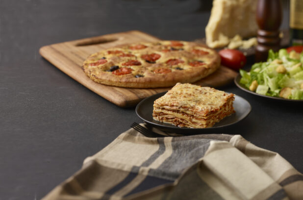 Slice of lasagna on a round black plate, focaccia, side salad, and other table items in background, dark background