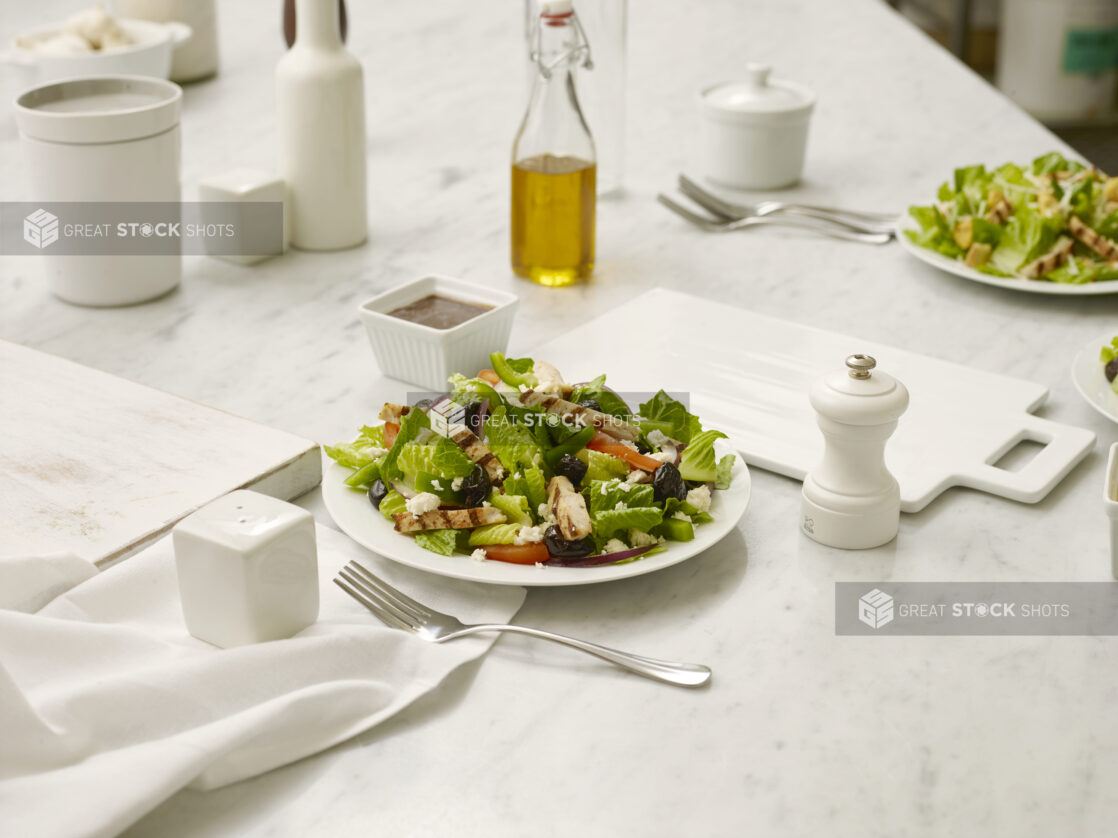 Greek Salad with grilled chicken in a white bowl, on a white marble table with white cutting boards, salt shaker, pepper grinder and a fork all in the foreground