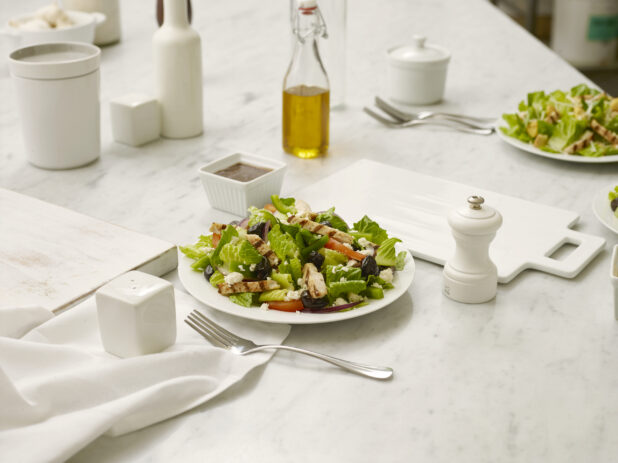 Greek Salad with grilled chicken in a white bowl, on a white marble table with white cutting boards, salt shaker, pepper grinder and a fork all in the foreground