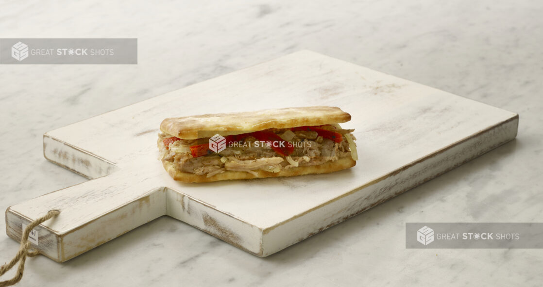 A single chicken panini/hoagie sandwich with red peppers and onions on a white cutting board on a white marble table