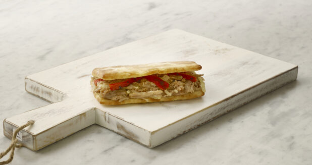 A single chicken panini/hoagie sandwich with red peppers and onions on a white cutting board on a white marble table