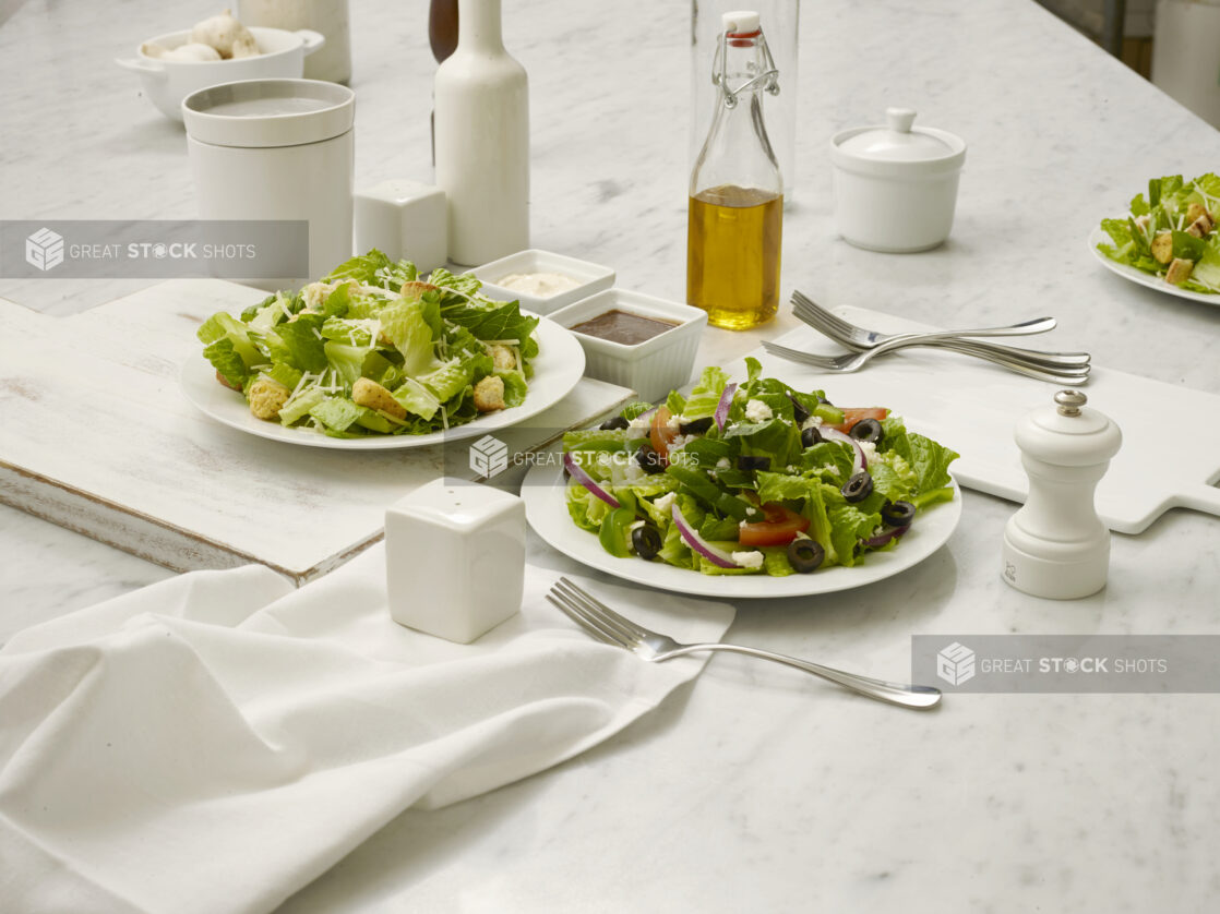 Caesar salad and Greek Salad in white bowls, on white cutting board and white marble table with accompanying tabletop items ie salt and pepper, etc., elegant