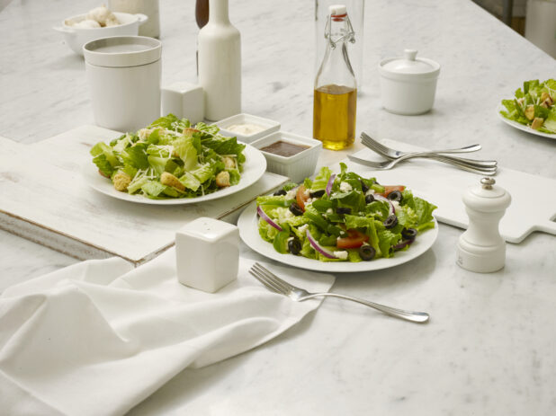Caesar salad and Greek Salad in white bowls, on white cutting board and white marble table with accompanying tabletop items ie salt and pepper, etc., elegant