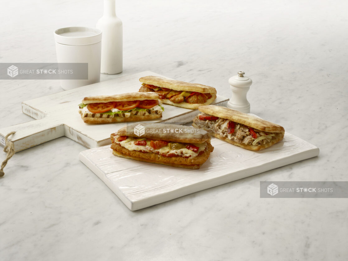 Grilled chicken, chicken parmesan, meatball and chicken shawarma sandwiches/hoagies on white cutting boards on a white marble table