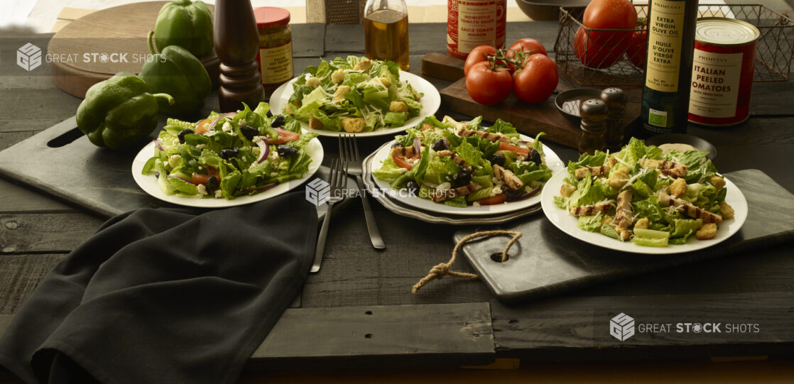 Two Caesar salads and two Greek salads in white bowls, one of each with grilled chicken and one without, on a black tabletop, black background