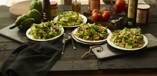 Two Caesar salads and two Greek salads in white bowls, one of each with grilled chicken and one without, on a black tabletop, black background