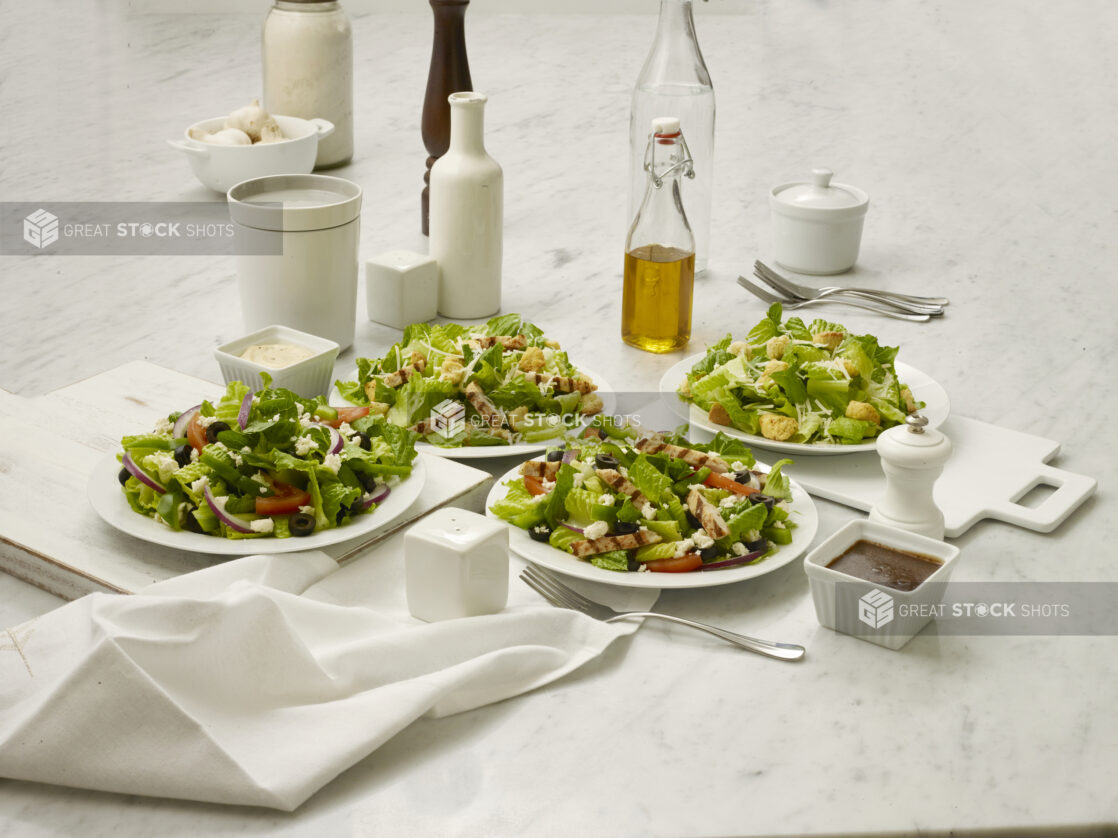 Two Caesar salads and two Greek salads in white bowls, one of each with grilled chicken and one without, on a white marble table top, white background