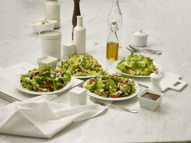 Two Caesar salads and two Greek salads in white bowls, one of each with grilled chicken and one without, on a white marble table top, white background