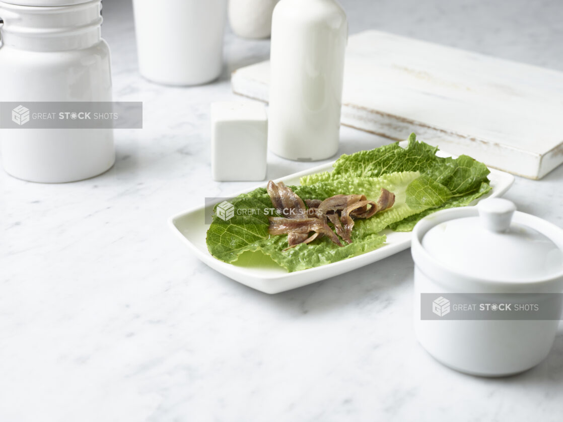 Anchovy fillets on a bed of baby romaine lettuce on a small square white plate in a white table setting