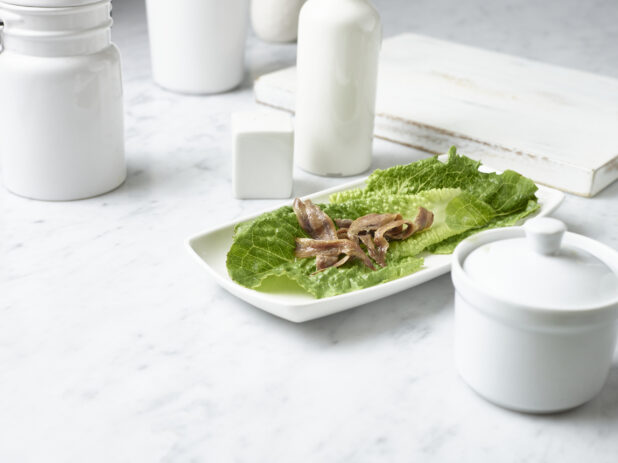 Anchovy fillets on a bed of baby romaine lettuce on a small square white plate in a white table setting