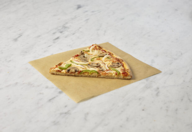 Classic Veggie Deluxe pizza slice on a square of parchment on a white marble background