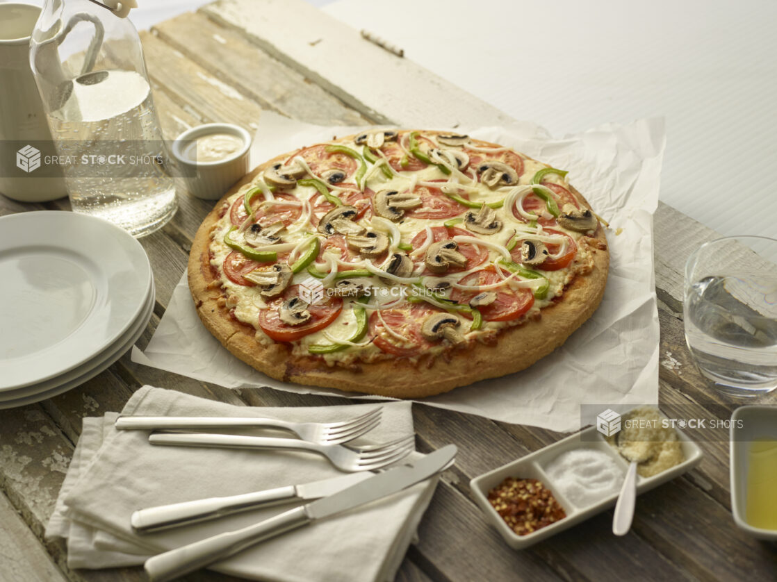 Whole Vegetarian deluxe pizza on parchment paper with utensils, napkins, condiments, and a carafe of water on a white wood table